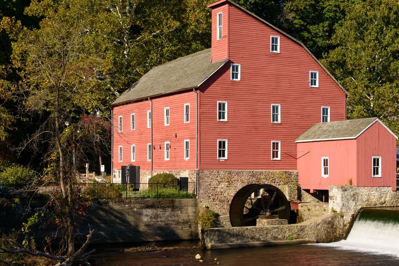 The Red Mill in Clinton, a red four-story mill with a waterwheel, beside a dam in autumn