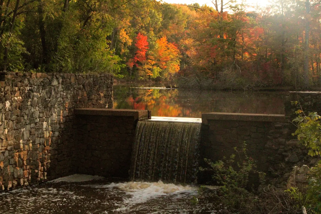A stone dam spilling water from Davidson's Mill Pond, with autumn trees reflected behind it