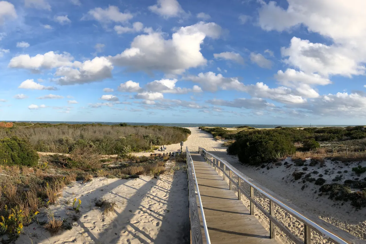 A boardwalk path through grassy dunes to the ocean at Barnegat Light, under scattered clouds